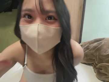 Media: Video of an Asian woman with long black hair, wearing a beige mask, white tank top, and a brown shirt, indoors, with a green wall and a brown cushion in the background.