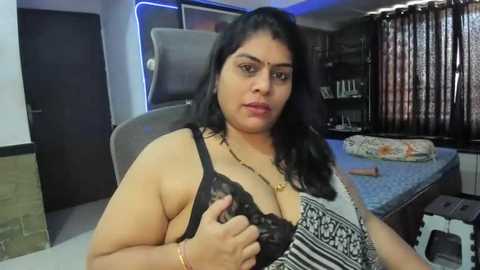 Media: Video of a plus-size woman with medium skin tone and black hair, wearing a black and white patterned bra, sitting on a chair in a dimly lit bedroom with a bed and curtains in the background.