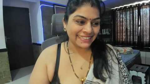 Media: A video of a smiling Indian woman with medium skin tone, wearing a black tank top, gold necklace, and bindi. She has long black hair and is in a modern, dimly-lit living room with a couch, curtains, and shelves.