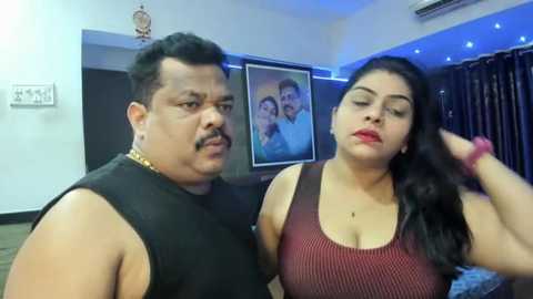 Media: Video of a middle-aged man and woman in a modern living room. He has a mustache, wearing a black tank top. She has long black hair, red lipstick, and a maroon ribbed top.