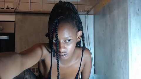 Media: Video of a young Black woman with braided hair, wearing a black spaghetti-strap top, in a dimly lit, concrete-walled kitchen. She appears to be taking a selfie.