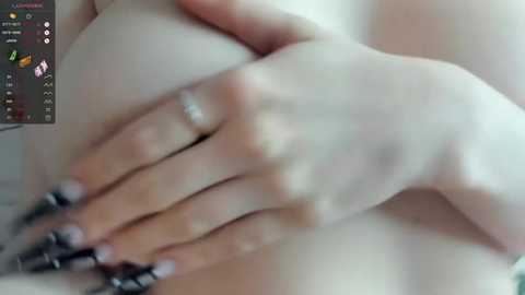 Media: A close-up video of a person's hand with neatly manicured nails, holding a breast. The image is slightly blurred, showing a soft focus. The background is a muted, out-of-focus setting.