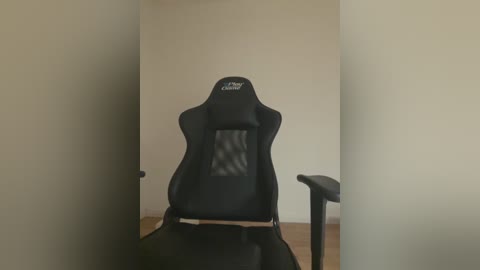 Media: A video of a black gaming chair with a high backrest and mesh fabric, set against a plain beige wall, positioned centrally in a minimalist room with wooden flooring.