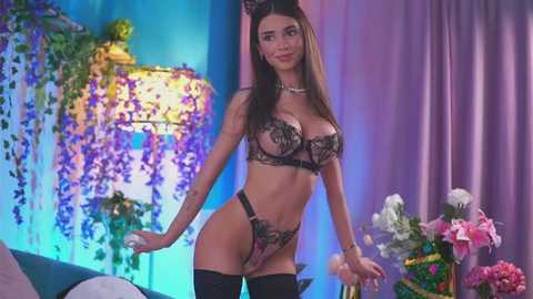 Media: Video of a slender, light-skinned woman with long, dark hair in black lingerie, standing in a vibrant, floral-themed room with purple curtains, a hanging purple blossom garland, and a pink floral arrangement.