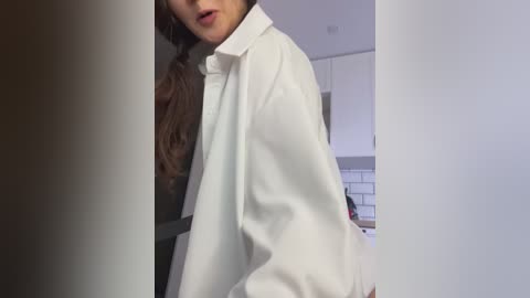 Media: Video of a young woman with long brown hair, wearing an oversized white button-down shirt, standing in a modern kitchen with white cabinets and subway tiles.