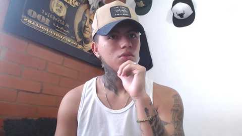 Media: Video of a young man with light skin and short black hair, wearing a tan baseball cap, white tank top, and black wristwatch. He's in a room with a brick wall and framed poster.