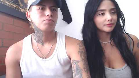 Media: Video of a young, tattooed man in a white tank top and baseball cap standing next to a young woman with long black hair in a gray tank top.