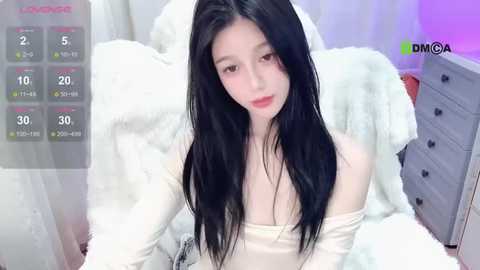 Media: A video of a young Asian woman with long black hair and fair skin, wearing a cream off-shoulder top, sitting on a white furry blanket in a bedroom with a gray dresser and a purple light.