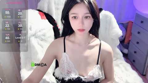 Media: Video of a young Asian woman with fair skin and long black hair, wearing a revealing white lace bra with black straps, sitting on a fluffy white blanket.