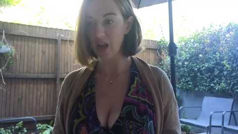 Media: Video of a fair-skinned woman with shoulder-length brown hair, wearing a low-cut, colorful-patterned top, standing in a backyard with wooden fence and greenery, mouth open as if mid-sentence.