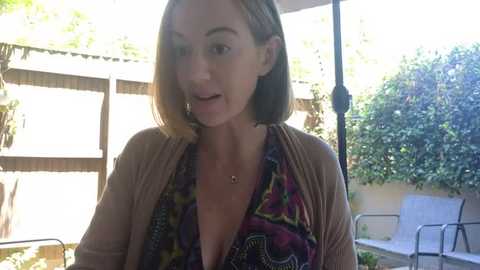 Media: Video of a Caucasian woman with shoulder-length brown hair, wearing a low-cut, patterned blouse and a beige cardigan, speaking outdoors in a sunny garden.