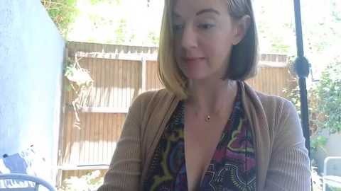 Media: Video of a smiling Caucasian woman with shoulder-length blonde hair, wearing a low-cut colorful patterned top and a beige cardigan, standing outdoors near a light blue wall and wooden fence.