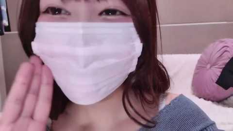 A video of a young woman with fair skin and long, dark brown hair, wearing a white surgical mask and a blue off-shoulder top. She is touching her face with her left hand.