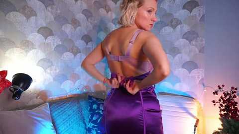 Media: Video of a blonde woman with fair skin in a purple satin dress, adjusting a bra strap, against a textured, circular-patterned wallpaper.