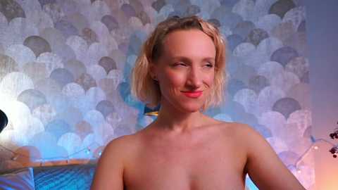 Media: Video of a smiling, fair-skinned, topless woman with blonde, shoulder-length hair. She has a slender physique and small breasts. Background features a modern, abstract wallpaper with large, circular patterns in blue, silver, and white.