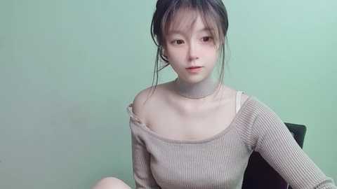 Media: Video of a young Asian woman with fair skin, wearing a beige ribbed sweater, sitting against a pale green wall. She has long, dark hair tied back and is looking directly at the camera.