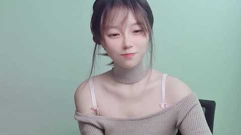 Media: Video of a young Asian woman with fair skin, dark hair, and bangs, wearing a beige off-shoulder sweater, pale pink bra straps, and a neutral background.