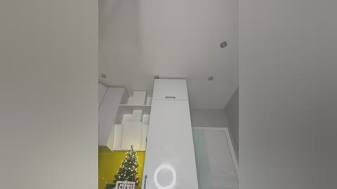 Media: A video of a modern, minimalist bathroom with white walls, a white sink, a small white cabinet, a yellow shelf, and a Christmas tree with white lights and ornaments on the left.