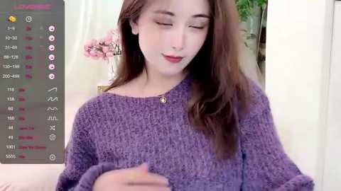 Media: A video of a young Asian woman with fair skin and long, straight brown hair, wearing a cozy, textured purple sweater. She appears relaxed, smiling gently, and holding a phone in her hands. The background features a light-colored room with a green plant and pink flowers.