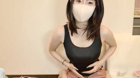 Media: Video of a young East Asian woman with fair skin and shoulder-length black hair, wearing a black tank top and beige face mask, seated indoors, hands on her stomach.