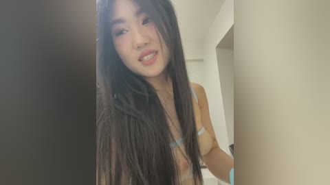 Media: Video of a young Asian woman with long, straight black hair, wearing a white bra, standing in a modern, minimalist bathroom with white walls.