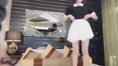 Media: Video of a slender, light-skinned woman in a black-and-white maid outfit, standing in a modern bedroom with a striped headboard, a decorative mirror, and beige pillows.