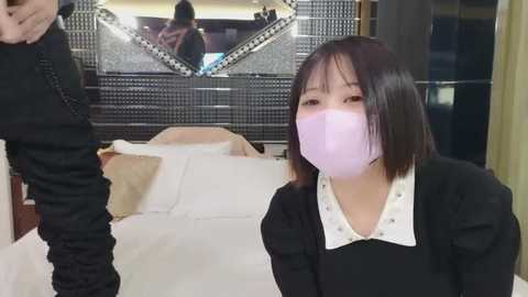 Media: Video of an Asian woman with straight black hair, wearing a black dress with white collar and a pink face mask, sitting on a white sofa. Background includes a mirror and a person in the reflection.