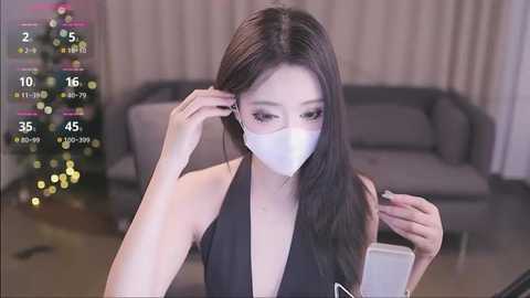 Media: A video of an Asian woman with long black hair, wearing a white face mask and a black halter top, adjusting her earphones in a modern living room with a Christmas tree and gray sofa.