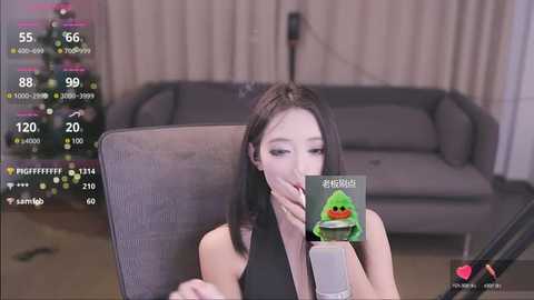 Media: A video of an Asian woman with long black hair, wearing a black tank top, holding a green microphone with a frog design. She is seated in a modern living room with a gray couch and a Christmas tree in the background.