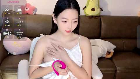 Media: A young East Asian woman with long black hair, slender build, and small breasts, wearing a white lace camisole, holds a pink vibrator on a brown couch.