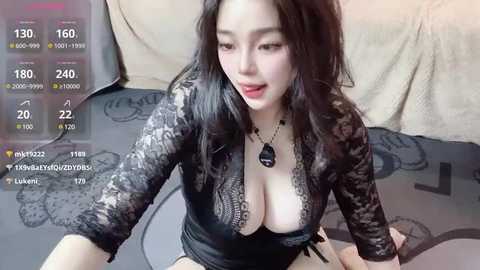 Media: A video of a young Asian woman with long black hair, wearing a black lace top that reveals ample cleavage, sitting on a bed. The background shows a beige blanket and a recording screen displaying viewer metrics.