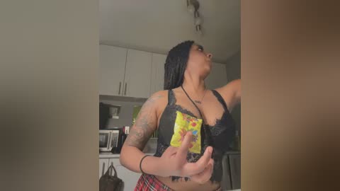 Media: Video of a plus-sized woman with medium-dark skin and black hair in a black lace bra, holding a yellow box, in a modern kitchen with white cabinets.