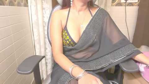 Media: Video of a woman in a semi-transparent black sari, revealing a yellow and black patterned bra, sitting in a modern office chair with a beige tiled wall and patterned curtains in the background.