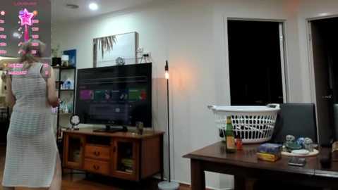 Media: A video of a cozy living room with a woman in a white dress watching TV, a laundry basket, and a wooden TV stand.