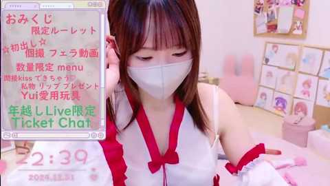 Media: A video of an Asian woman with straight brown hair, wearing a white mask, red dress, and pink ribbons, sitting on a bed in a room with pink walls and a poster. Text in Japanese overlays the image.