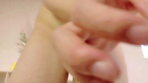 A blurry video of a person's hand, fingers intertwined, with a hint of a bare lower torso, set against a light-colored, indistinct background.