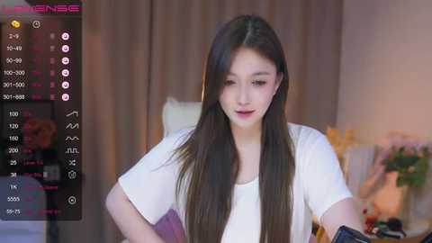 Media: Video of an Asian woman with long black hair, fair skin, and wearing a white t-shirt, captured during a live stream on a phone. Background shows blurred indoor setting with curtains and a table.