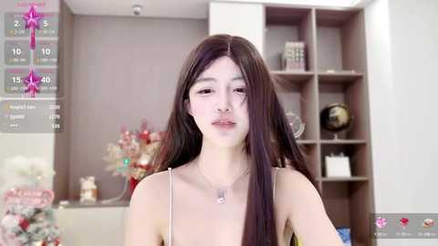 Media: A video of an Asian woman with long black hair, wearing a beige spaghetti-strap dress, standing in a modern, minimally decorated room with a gray background.