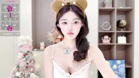 Media: Video of a young Asian woman with fair skin, long dark hair, and a teddy bear headband. She wears a white lace camisole, with a small badge on her chest. Background shows a cozy room with a decorated Christmas tree, shelves, and a blue screen.