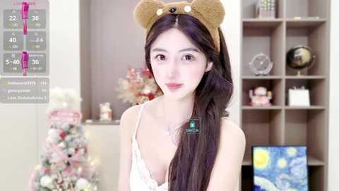 Media: Video of a young Asian woman with fair skin and long black hair, wearing a teddy bear headband and a white lace bralette, standing in a cozy, Christmas-decorated room with a Starry Night painting.