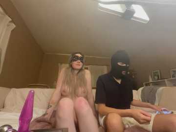 Media: A video depicts a nude woman with long blonde hair wearing a black mask, sitting next to a man in a black mask, both holding sex toys on a beige couch in a dimly lit room.