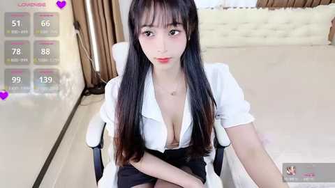 Media: A video of an East Asian woman with long black hair, wearing a white blouse revealing cleavage, sitting on a white chair in a modern room.