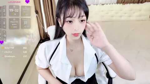 Media: Video of an Asian woman with long black hair, wearing a white blouse unbuttoned to reveal cleavage, sitting in a modern bedroom.