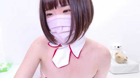 Media: Video of an East Asian woman with straight, shoulder-length dark hair, wearing a white nurse's mask and uniform with a white collar and red trim, standing in a bright, minimalist room with a teddy bear.