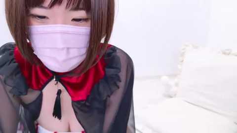 Media: Video of a woman with a white face mask, short brown hair, and a black sheer blouse with red ruffled collar and tassel, kneeling on a white bed, looking down.