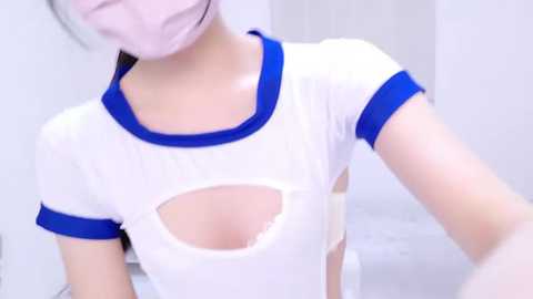 Media: Video of an Asian woman with a pale complexion wearing a white, see-through T-shirt with blue trim, revealing a large cleavage cutout, and a pink surgical mask. Background is a white, sterile environment.