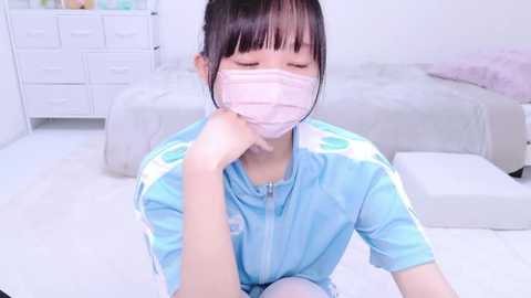 Media: Video of an Asian woman with long black hair, wearing a blue jacket, white mask, and white gloves, kneeling on a white floor in a minimalist bedroom with a bed and white dresser in the background.