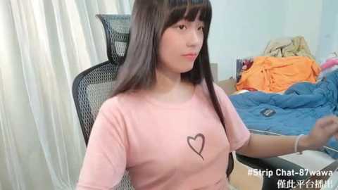 Media: Video of a young East Asian woman with long black hair, wearing a pink t-shirt with a heart graphic, sitting in a black office chair in a messy bedroom with a bed and orange blanket.