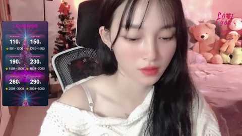 Media: Video of an East Asian woman with fair skin, long black hair, and a white off-shoulder sweater, sitting in a cozy room with a Christmas tree and stuffed animals.