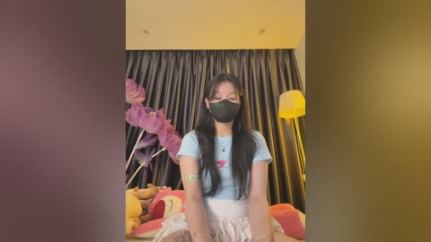 Media: Video of an East Asian woman with long black hair, wearing a light blue T-shirt and a face mask, seated in a room with a plush teddy bear, purple flowers, and a yellow lamp in the background.